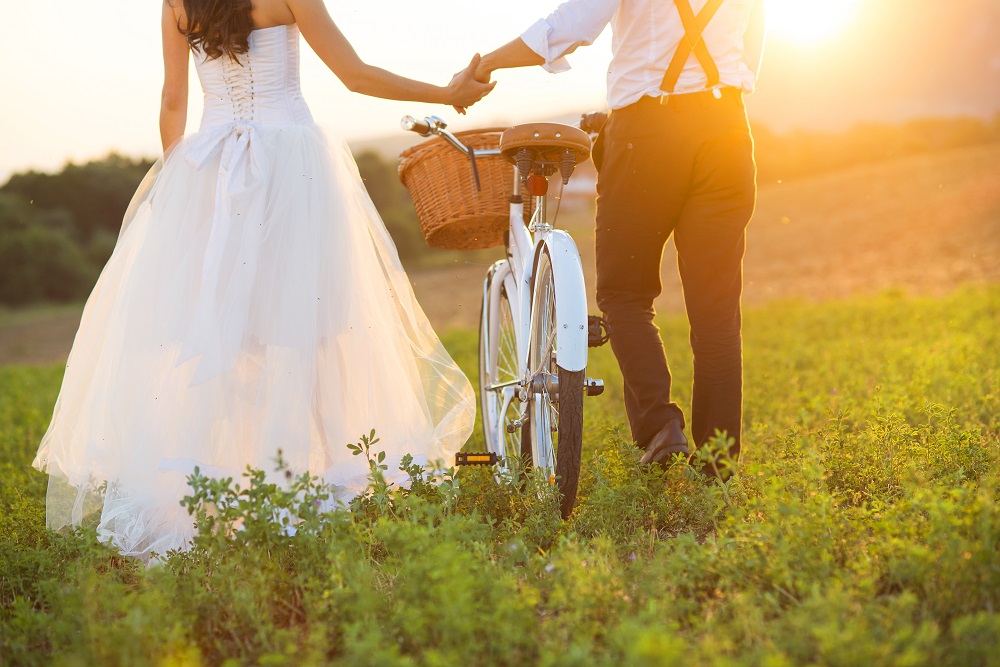 Para młoda (w strojach ślubnych) idzie przez łąkę z rowerem