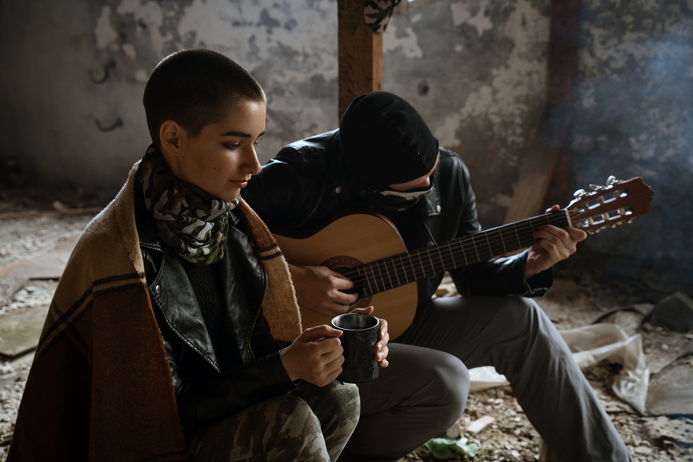 Krótko obcięta kobieta trzymająca kubek i mężczyzna grający na gitarze w zniszczonym, obdrapanym wnętrzu
